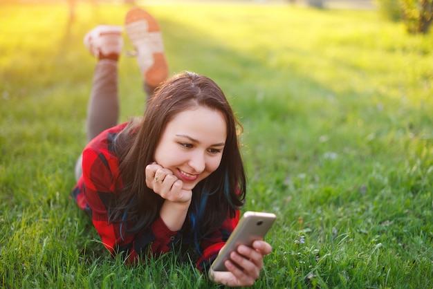 Photo portrait of a smiling woman lying on green grass and using smartphone outdoors