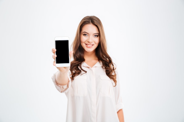 Portrait of a smiling woman showing blank smartphone screen isolated on a white wall