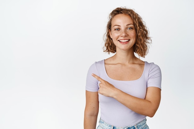 Portrait of smiling woman showing sale, pointing finger left, advertising product, standing in tshirt over white background