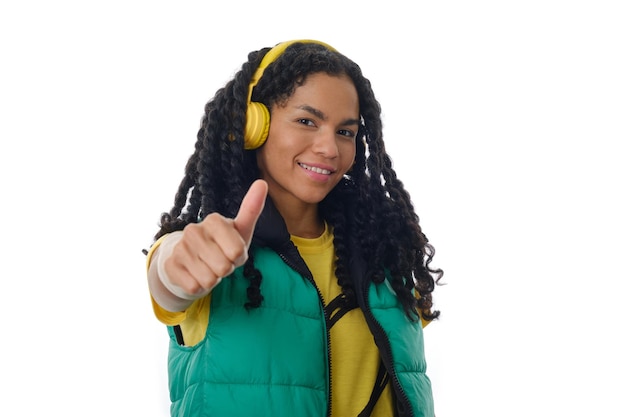 Foto ritratto di una donna sorridente che mostra pollice in su mentre ascolta la musica con le cuffie su uno sfondo isolato.