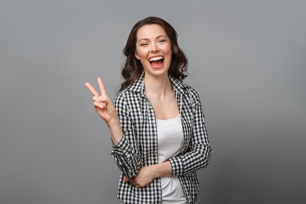 Ritratto di una donna sorridente che mostra un segno di vittoria e guarda in una telecamera isolata su un grigio