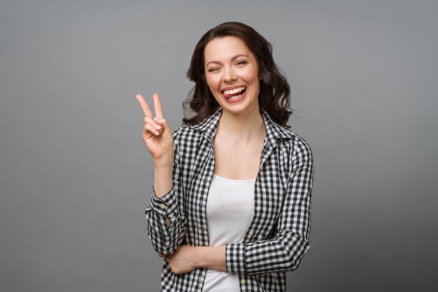 Portrait of a smiling woman showing a victory sign and looking into a camera isolated on a gray