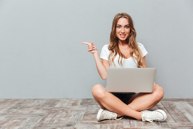 Portrait of a smiling woman sitting on the floor with laptop and pointing finger away isolated on gray wall