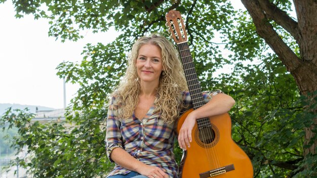 Photo portrait of smiling woman sitting with guitar against trees