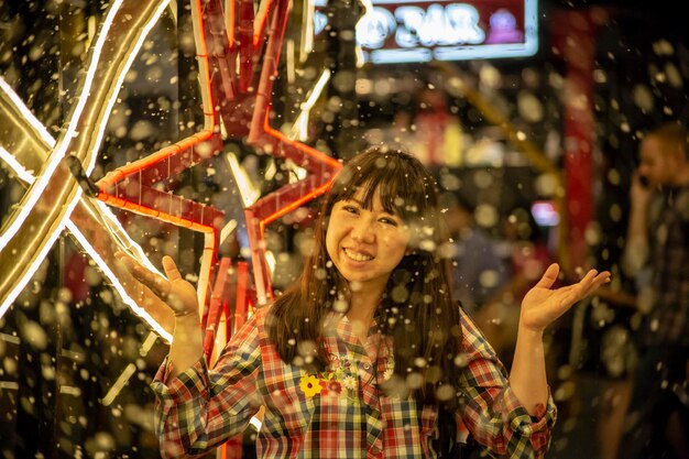 Foto ritratto di una donna sorridente in piedi davanti a una decorazione illuminata durante la nevicata notturna