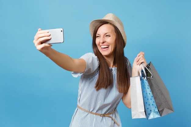 Portrait smiling woman in summer dress, straw hat holding packages bags with purchases after shopping doing selfie shot on mobile phone isolated on blue pastel background. Copy space for advertisement