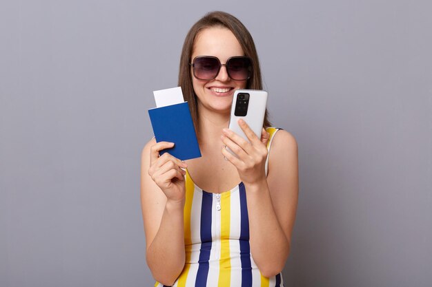 Портрет улыбающейся женщины-туриста с мобильным телефоном и паспортом, которая хочет отправиться в летнее путешествие за границу, пытается зарезервировать отель в полосатом купальнике, изолированном на сером фоне