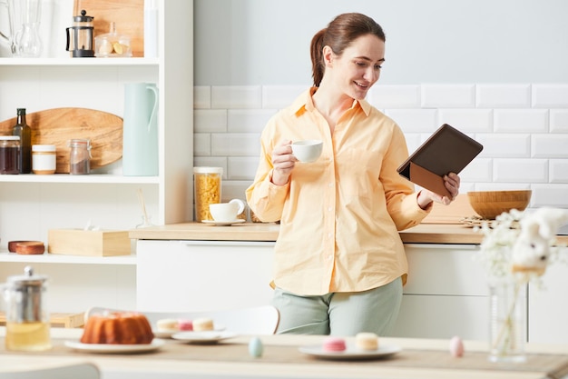 Портрет улыбающейся женщины, использующей цифровой планшет на кухне, наслаждаясь завтраком в утреннем пространстве для копирования