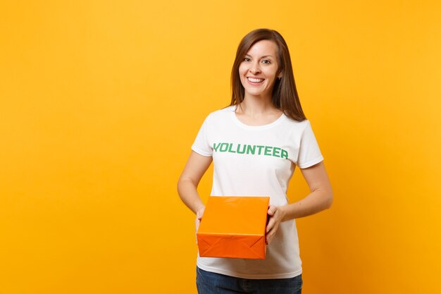 Portrait of smiling woman in white t-shirt with written inscription green title volunteer hold orange cardboard box isolated on yellow background. Voluntary free assistance help, charity grace concept