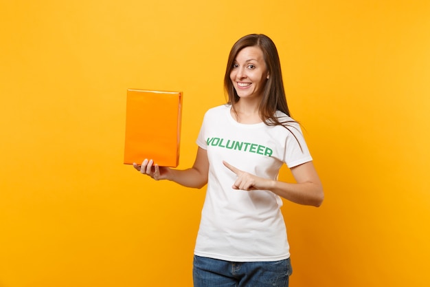 Ritratto di donna sorridente in t-shirt bianca con iscrizione scritta titolo verde volontario tenere scatola di cartone arancione isolata su sfondo giallo. aiuto volontario di assistenza gratuita, concetto di grazia di beneficenza