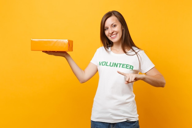 Ritratto di donna sorridente in t-shirt bianca con iscrizione scritta titolo verde volontario tenere scatola di cartone arancione isolata su sfondo giallo. aiuto volontario di assistenza gratuita, concetto di grazia di beneficenza