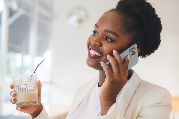 Portrait of smiling woman with afro hairstyle talking phone and looking at out of the window while sitting in cafe Attractive millennial woman with cellular device Talking with friend