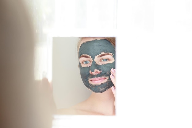 Portrait of a smiling woman with a black clay cosmetic mask on\
her face looks in the mirror self care at home