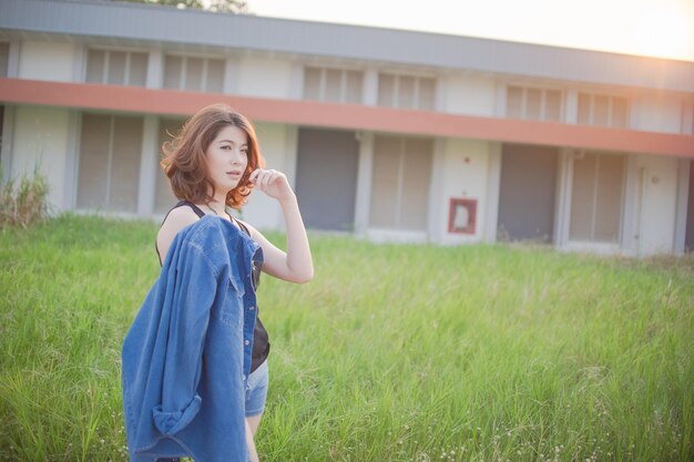 Portrait of smiling woman with denim jacket standing on field against building during sunset