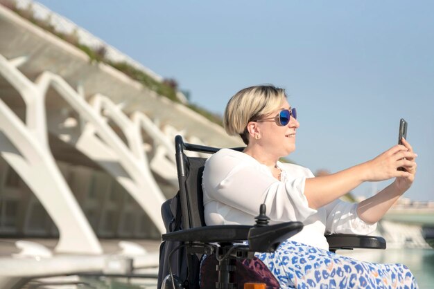 Foto ritratto di una donna sorridente con disabilità su una sedia a rotelle con occhiali da sole che scatta una foto selfie