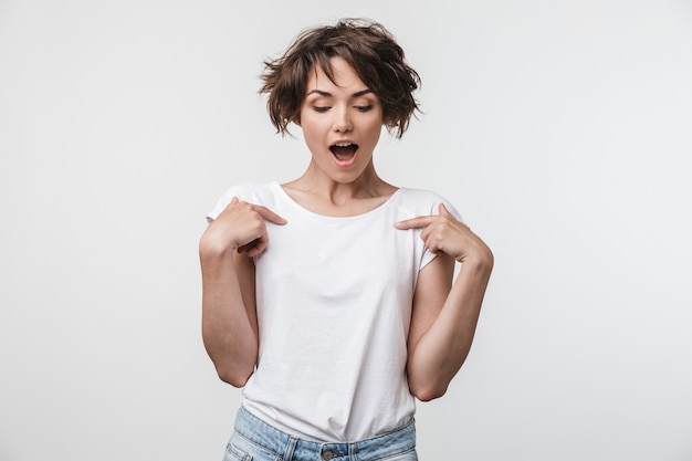 Portrait of smiling woman with short brown hair in basic t-shirt rejoicing and pointing fingers at herself isolated over white wall