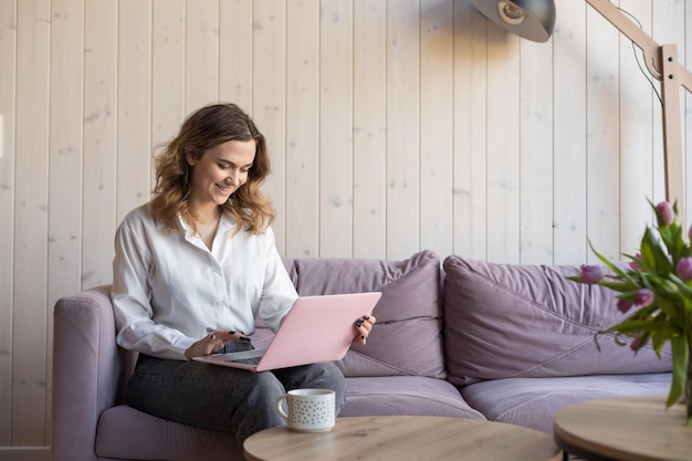 Portrait of smiling woman working on pink laptop sitting on\
soft violet sofa in casual clothes near