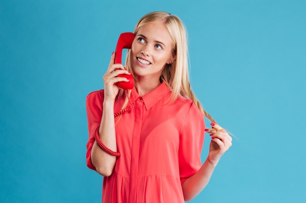Портрет улыбающейся удивленной женщины в красном платье, говорящей по мобильному телефону и смотрящей в сторону, изолированной на синей стене