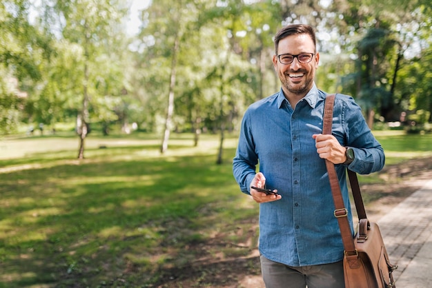 Portrait of smiling young adult man walking through the park using phone