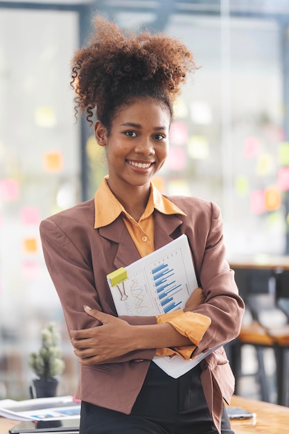 Portrait of smiling young african american businesswoman holding papaerwork standing in office