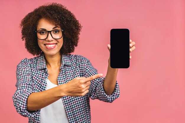 Portrait of a smiling young african american woman holding\
blank screen mobile phone and showing finger isolated over pink\
background