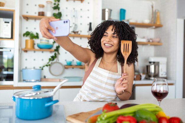 自宅のキッチンで料理をしながらスマートフォンで自撮りをする笑顔の若いアフリカ系アメリカ人女性のポートレート