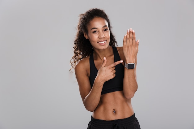 Portrait of a smiling young african sportswoman pointing at blank screen smart watch isolated over gray background