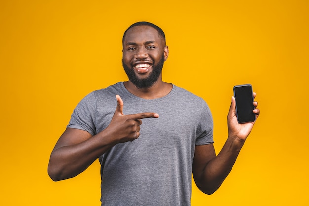 Il ritratto di giovane uomo afroamericano sorridente si è vestito in isolato casuale, indicando al telefono cellulare dello schermo in bianco.