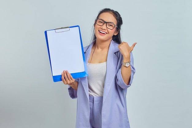 Portrait of smiling young Asian business woman showing blank clipboard and pointing to copy space with finger isolated on purple background