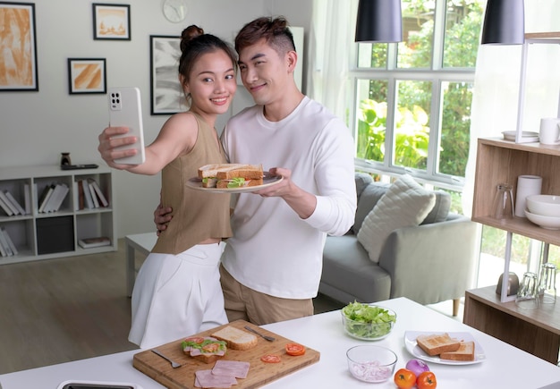 Portrait of a smiling young Asian couple taking a selfie while cooking together at the kitchen Happy young man and young woman are having fun together while prepare food Couple lifestyle concept