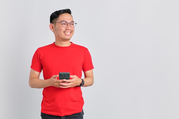 Portrait of smiling young Asian man holding mobile phone and looking aside at copy space isolated over white background