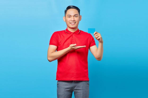 Ritratto di giovane uomo asiatico sorridente che mostra la carta di credito, offre un prodotto pubblicitario isolato su sfondo blu