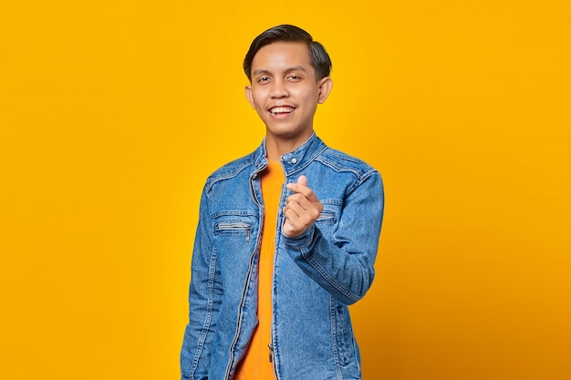 Портрет улыбающегося молодого азиатского человека, показывающего сердце пальцем на желтом фоне
