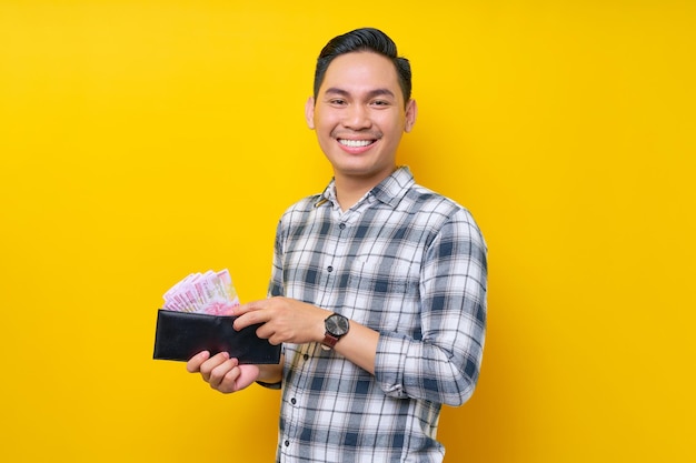 Portrait of smiling young Asian man wearing plaid shirt holding a wallet full of cash money in rupiah banknotes in hand isolated on yellow background people lifestyle concept
