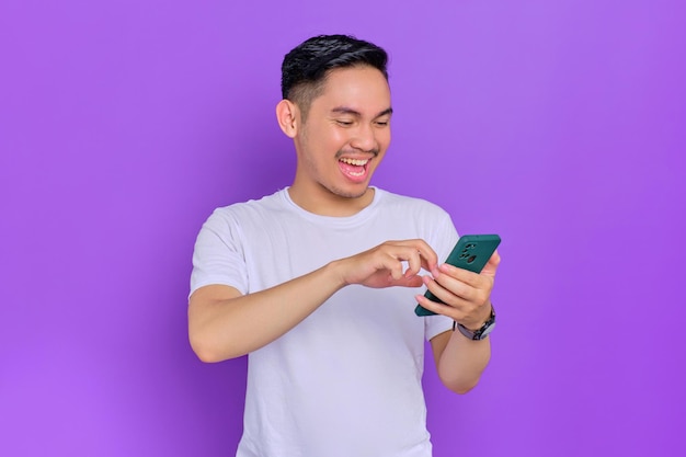 Portrait of smiling young Asian man in white tshirt using smartphone for messaging with friends isolated on purple background