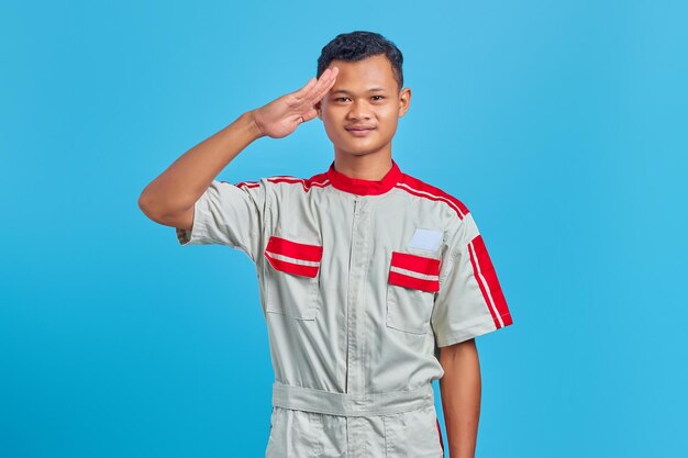 Portrait of smiling young Asian mechanic making salute sign with palm isolated on blue background