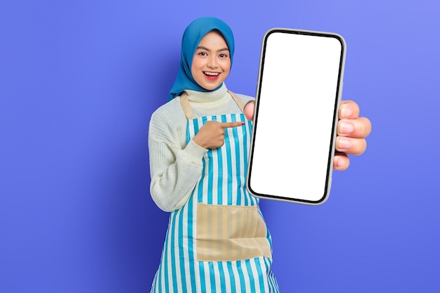 Portrait of smiling young asian muslim woman in 20s wearing hijab and apron pointing blank screen mobile phone with finger isolated over purple background people housewife muslim lifestyle concept