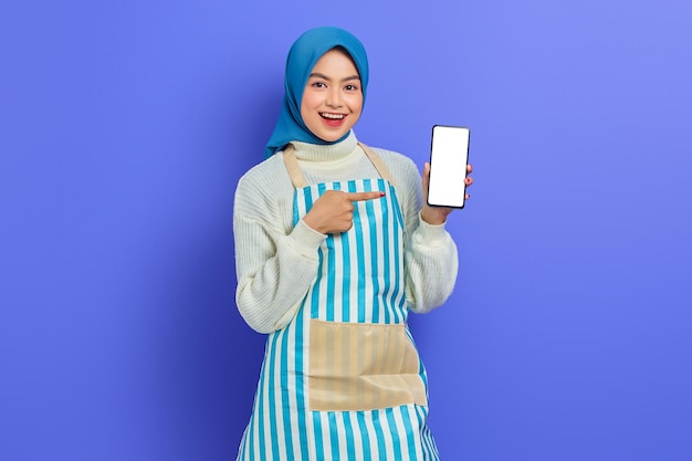 Portrait of smiling young asian muslim woman in 20s wearing hijab and apron pointing blank screen mobile phone with finger isolated over purple background. people housewife muslim lifestyle concept