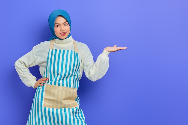 Portrait of smiling young asian muslim woman in hijab and\
striped apron pointing aside with hands while doing housework\
isolated on purple background people housewife muslim lifestyle\
concept