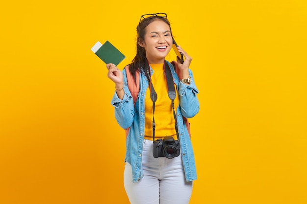 Portrait of smiling young Asian traveler holding boarding pass passport ticket and talking on smartphone isolated on yellow background. Passengers traveling on weekends. Air flight travel concept