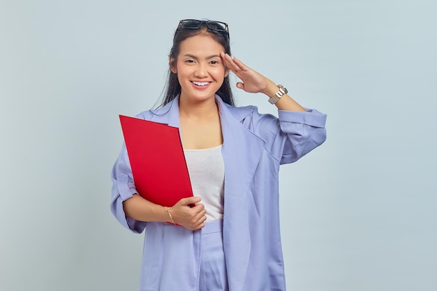 Ritratto di giovane donna asiatica sorridente che tiene la cartella dei documenti e che gesticola rispetto al superiore, pronta a fare il lavoro d'ufficio isolato su sfondo viola