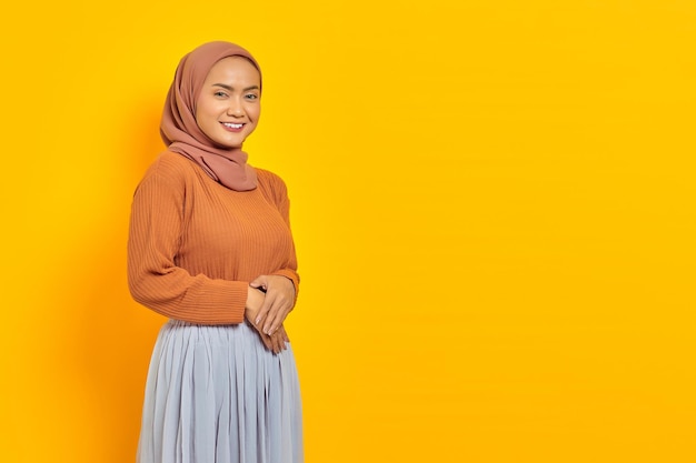 Portrait of smiling young Asian woman holding hands together and feels optimistic isolated on yellow background