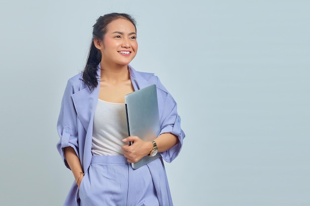 Портрет улыбающейся молодой азиатки, держащей ноутбук и смотрящей в сторону на белом фоне