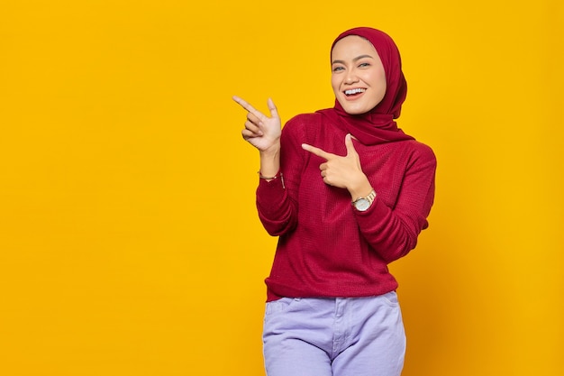 Portrait of smiling young Asian woman pointing finger at copy space on yellow background