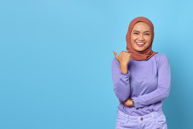 Portrait of smiling young Asian woman pointing a thumb at copy space on blue background