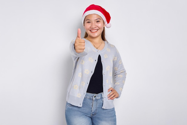 Portrait of smiling young Asian woman in Santa Claus hat showing thumbs up isolated over white background