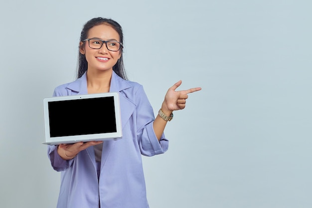 Portrait of smiling young Asian woman showing blank laptop screen and pointing to copy space with finger isolated on white background