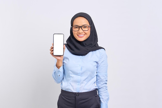 Portrait of smiling young Asian woman showing blank screen mobile phone and looking at camera isolated on white background