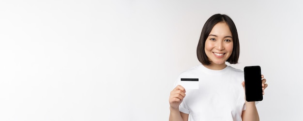 Portrait of smiling young asian woman showing mobile phone screen and credit card standing over whit