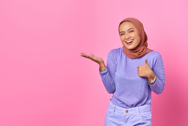 Portrait of smiling young Asian woman showing palm hands and thumbs up on pink background